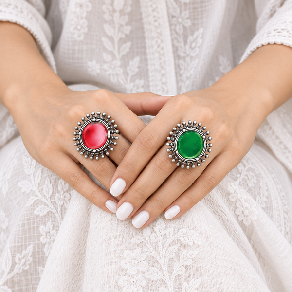 Hands wearing two oxidised 925 sterling silver statement rings with round red and green stones and beaded halos