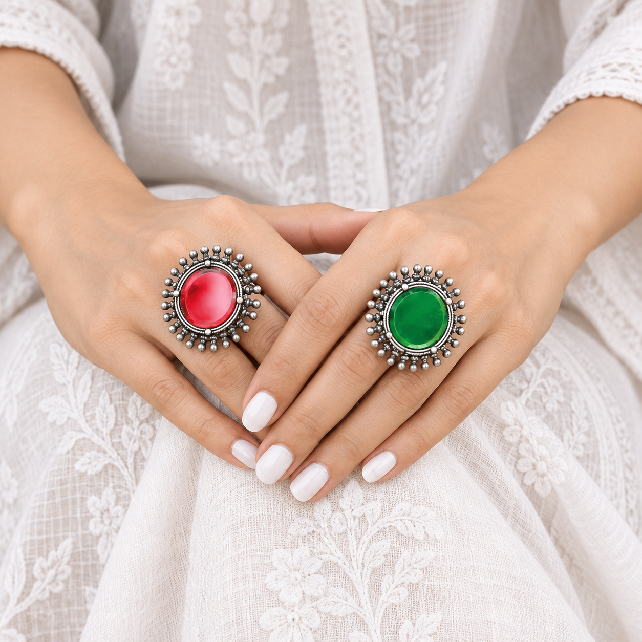 Hands wearing two oxidised 925 sterling silver statement rings with round red and green stones and beaded halos