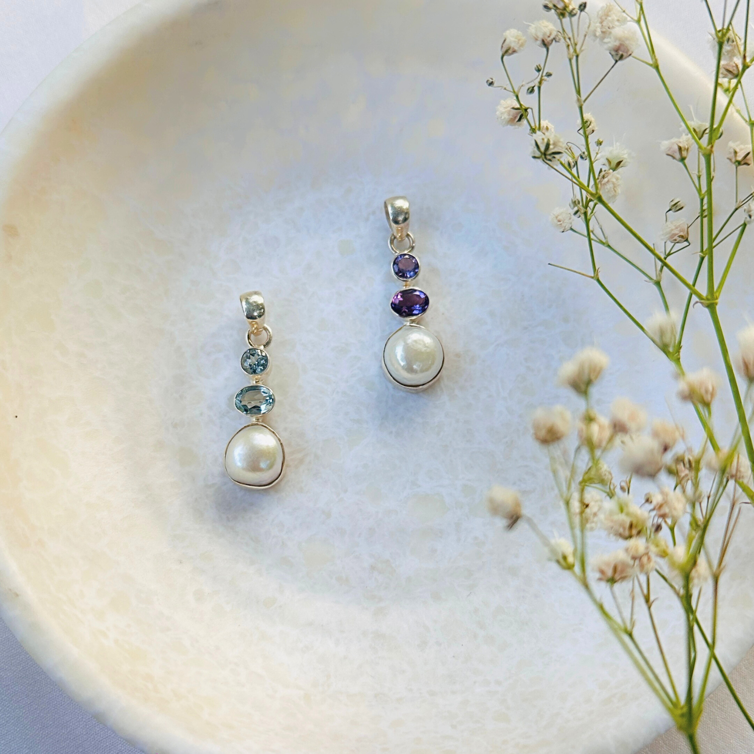 Two pearl pendants with gemstones on a textured surface with flowers.