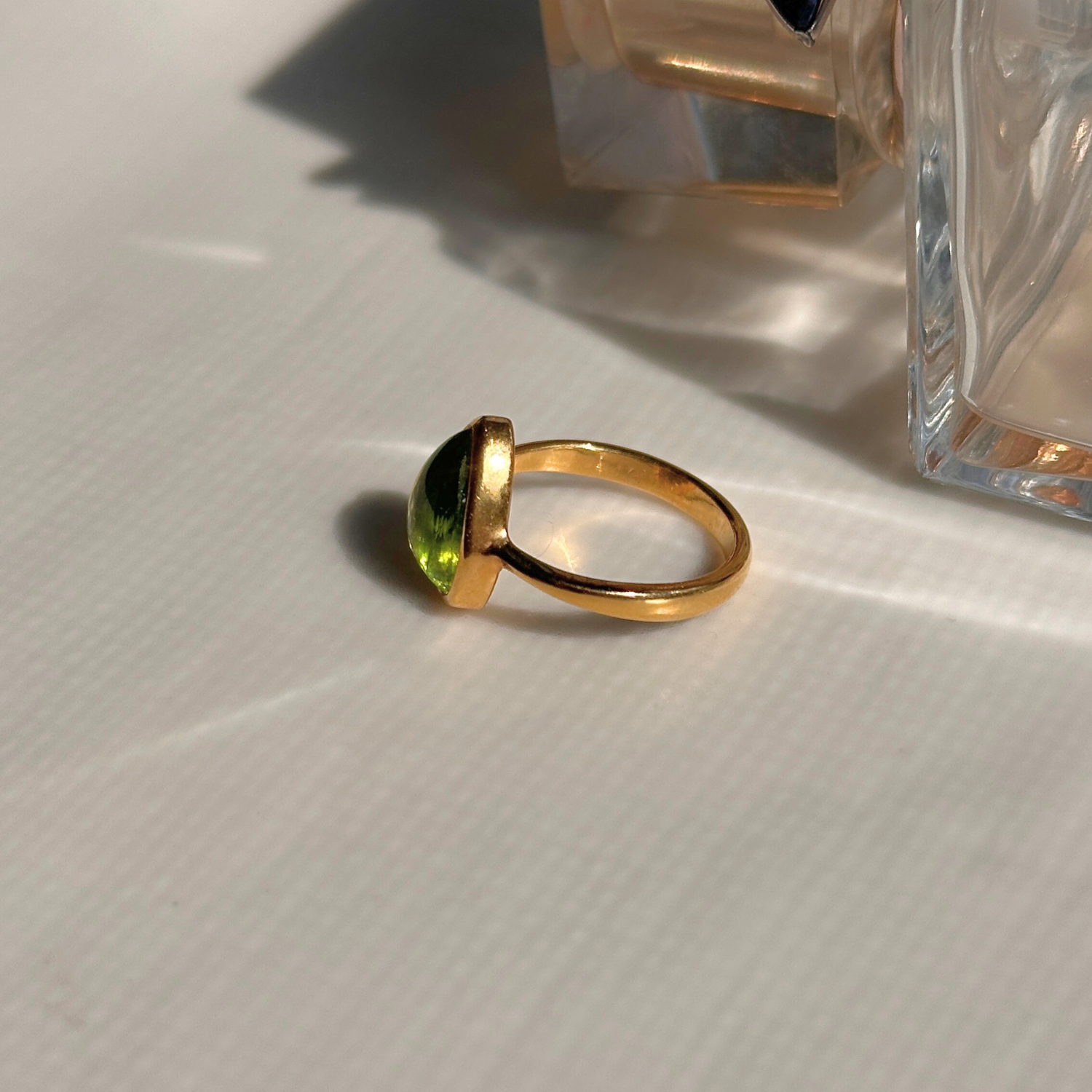 Gold ring with a green gemstone on a textured white surface with a clear glass container in the background.